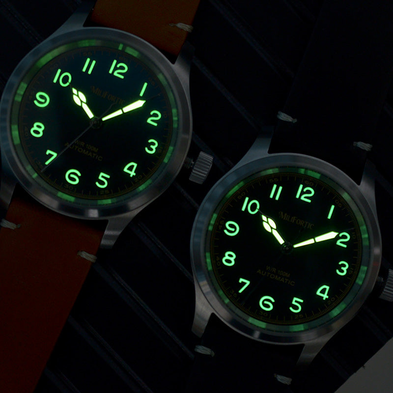 Two Murph watches lying flat on a table in a dim setting, showing bright green lume