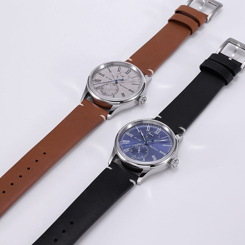 Two dress watches lying flat on a table, one with a blue dial and the other with a gray dial