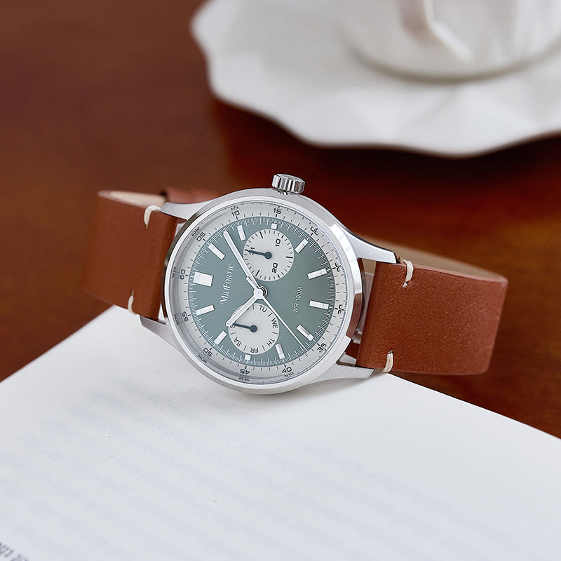 R078 watch with green dial, brown strap, angled on the side of a book on a table.